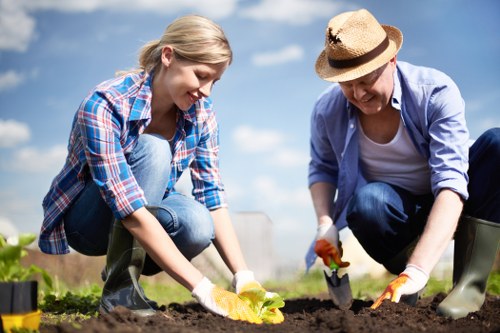 A young woman and an older woman are working together in a garden on a sunny day, both dressed casually with the woman in a blue plaid shirt and the older woman in a light blue jacket and straw hat. They are actively planting or tending to the dark, moist soil in a raised garden bed, with gardening gloves on their hands. The garden area features well-maintained flower beds and surrounds a lush, green outdoor space, set against a bright blue sky with some scattered clouds. The scene depicts outdoor gardening work suitable for lawn and garden care services, highlighting community involvement in landscape maintenance in Clapham, with visible gardening tools nearby. The lighting indicates clear weather, suggesting ideal conditions for planting or garden tending activities, which underscores the importance of professional gardening services offered by Landscapers Clapham.