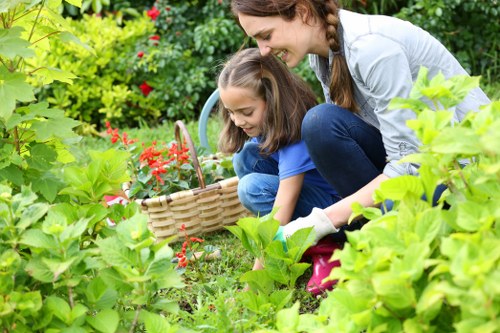 A woman and a young girl are gardening together in a lush, well-maintained backyard, surrounded by vibrant green foliage, bushes, and flowering plants. They are focused on harvesting or planting in a small flower bed bordered by neatly trimmed shrubs. The woman is crouching down and smiling, wearing casual outdoor clothing, while the girl, dressed in a blue top, is also actively engaged in gardening. Behind them, there is a variety of garden elements including a wicker basket filled with red flowers, and a serene outdoor environment with natural sunlight illuminating the scene. The garden has a mix of textured paving and grassy areas, offering an inviting outdoor space for leisure and landscaping activities, aligned with the services provided by Landscapers Clapham, specialists in gardening and outdoor maintenance in the Clapham area, inferred from the postcode SW4 in South London.