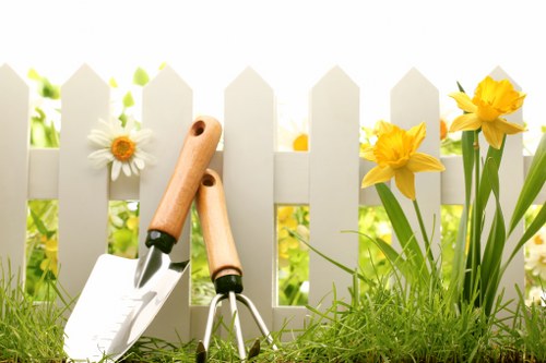 The image depicts a section of a garden enclosed by a white picket fence, with lush green grass in the foreground and flowering plants along the fence line. There are yellow daffodils and white daisies with orange centers growing beside the fence, adding natural colour and variety to the garden. In front of the fence, garden tools including a hand trowel and a small garden fork with wooden handles are planted into the soil, indicating ongoing planting or maintenance work. The environment suggests a well-kept outdoor space suitable for gardening services, with bright natural lighting suggesting a sunny day and a clear sky. The overall scene highlights the garden's landscaped layout, with neatly arranged flower beds and a healthy, vibrant lawn, ideal for sustaining or improving landscaping efforts in the Clapham area or similar suburban settings.