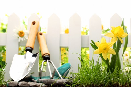 The image depicts a section of a well-maintained garden with a white picket fence in the background, characteristic of outdoor spaces in Clapham. In the foreground, there is a lush, green lawn with dense, evenly cut grass, bordered on one side by mature yellow daffodils in full bloom, showcasing bright yellow petals and green, strap-like leaves. The garden features a small flower bed area near the fence, with some white daisies and other small flowering plants visible. Resting on the soil near the flowers are garden tools, including a hand trowel with a wooden handle, a small garden fork, and gardening gloves, suggesting recent or ongoing gardening activities. The environment appears to be outdoors on a bright, sunny day, with natural light illuminating the scene and casting soft shadows, emphasizing the vibrant colours and textures of the plants and garden accessories. This scene exemplifies a typical landscaped outdoor space suitable for lawn care, gardening, and landscape maintenance services offered by Landscapers Clapham in the SW4 postcode area, highlighting the importance of proper lawn and garden management in residential settings.