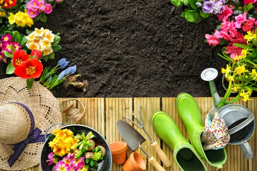 The image depicts a section of a well-maintained garden outdoor space, featuring a wooden deck border adjacent to a lawn area with rich, dark soil. On the deck, there are various gardening tools including a pair of green rubber gloves, a small trowel, a hand rake, and empty flower pots. To the left, there is a wide-brimmed straw hat with a purple ribbon, and nearby, a black planting container filled with colorful blooming flowers such as primroses and pansies, adding vibrant yellow, pink, and purple hues. Surrounding the deck, the garden is decorated with flower beds containing a variety of flowering plants, with some blooms spilling into the scene's edges and demonstrating natural diversity in colour and size. The garden environment appears bright and sunny, with no visible signs of rain or overcast weather, suggesting good conditions for outdoor gardening. This setting exemplifies a typical backyard garden area suitable for gardening services offered by companies like Landscapers Clapham, incorporating lawn maintenance and flower planting in a neat, accessible outdoor space near Clapham, London.