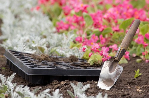 A close-up of a gardening scene in a backyard garden in Clapham, featuring a small shovel with a wooden handle planted in the dark, moist soil. Surrounding the planting area, there are various flowering plants with vibrant pink and white blossoms, creating a colourful border. The soil appears freshly turned, indicating recent planting or garden maintenance. In the background, there is a black plastic seed tray partially filled with soil, ready for sowing seeds or transplanting seedlings. The garden has a well-organized layout with flower beds delineated by soil borders, and the natural outdoor environment is illuminated by natural daylight, suggesting a clear, calm day. This scene reflects typical garden maintenance activities, such as planting, soil preparation, and flower bed enhancement, aligning with gardening and landscaping services provided by companies like Landscapers Clapham, based in South West London.