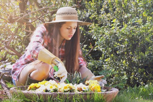 A young woman wearing a wide-brimmed straw hat, a checkered shirt, and gardening gloves is kneeling in a lush, green garden. She is tending to a flower bed bordered by soil and grass, carefully planting or arranging vibrant yellow and white flowers. The garden features a well-maintained lawn with dense greenery and shrubs in the background, indicating a cultivated outdoor space. Sunlight filters through the foliage, illuminating the scene with natural light, suggesting a mild, sunny day. This outdoor environment showcases typical features of a landscaped garden in Clapham, with a focus on floral planting and lawn care, reflecting professional gardening and landscaping practices common in the area. Landscaping services by Landscapers Clapham often include flower bed planting, lawn maintenance, and shrub trimming to enhance garden aesthetics and functionality.