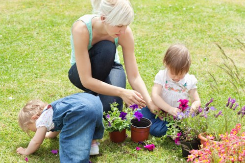 A woman with short blonde hair, wearing a light blue sleeveless top and dark jeans, kneels on a well-maintained grassy lawn in a garden, tending to potted plants and flowers. Beside her, a young girl with light brown hair, dressed in a white top with a colorful pattern, is seated on the grass, carefully examining or tending to bright pink and purple flowers in a black container. Another young child, with blond hair and wearing blue overalls, is crouched on the grass, reaching toward the ground near the plants. The garden area features lush, green grass, and vibrant, colorful blossoms suggest a well-kept outdoor space. The background includes a simple, tidy garden environment, with natural lighting indicating a clear, sunny day. This scene highlights outdoor gardening activity suitable for family participation, emphasizing maintenance of flower beds and potted plants, consistent with the gardening services offered by Landscapers Clapham in the local Clapham area, post code SW4.