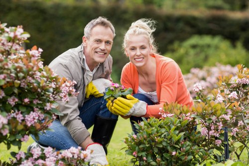 A man and a woman, both smiling, are kneeling in a well-maintained garden with a variety of blooming pink and white flowers and green foliage in the foreground. The man has short grey hair and wears a grey shirt, while the woman has blonde hair tied back and wears a coral jacket with yellow gardening gloves. Behind them, the garden features lush, dense greenery with a hedge and a distant tree line under a partly cloudy sky, suggesting a mild spring or summer day in Clapham. The scene depicts outdoor gardening activity and the care of flowering shrubs and plants, reflecting professional gardening and landscaping services provided by Landscapers Clapham, aligning with their gardening service offerings on the Clapham patio repair solutions page.